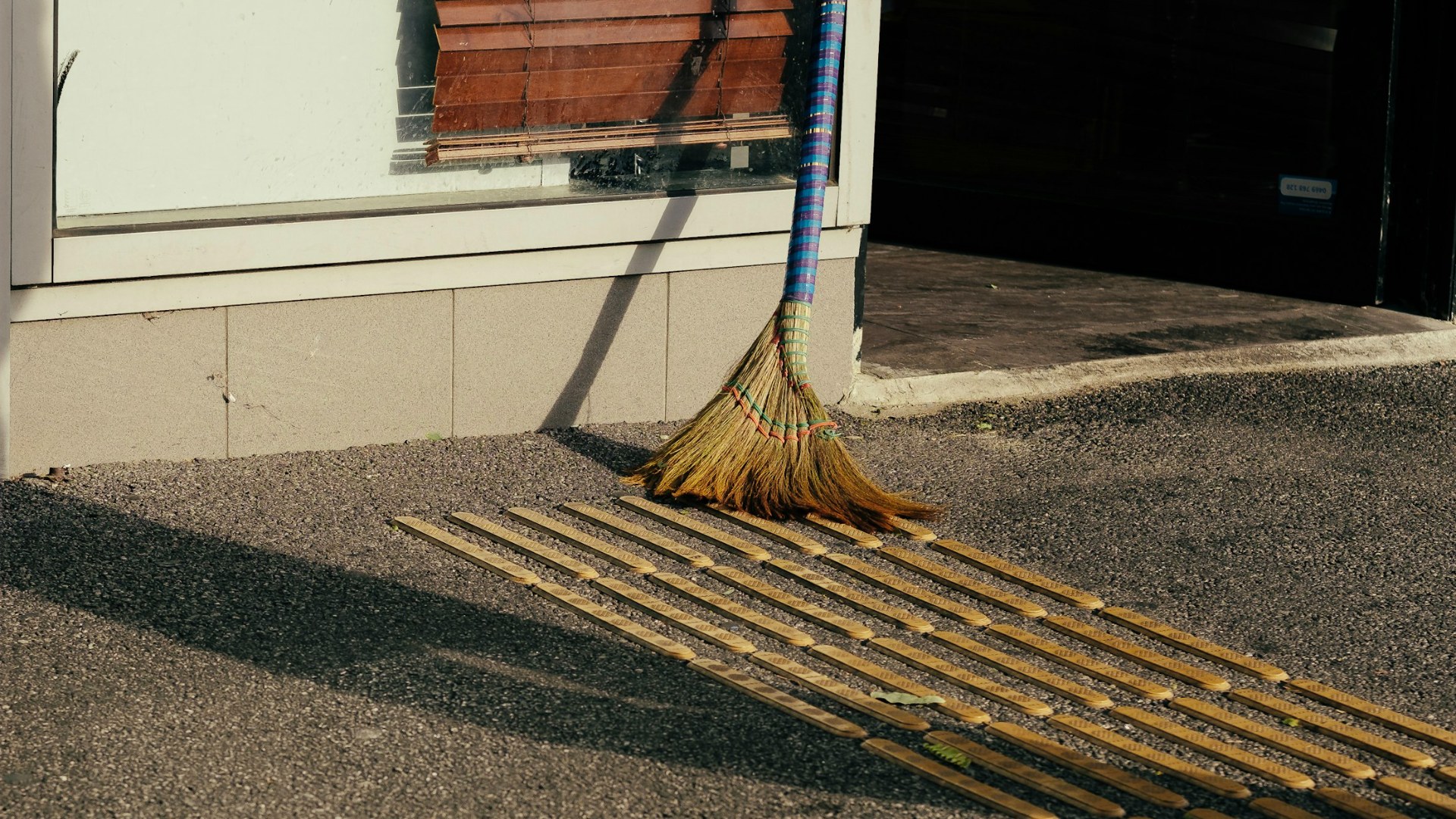 A traditional straw broom leaning against a shopfront in morning light, beside yellow tactile paving