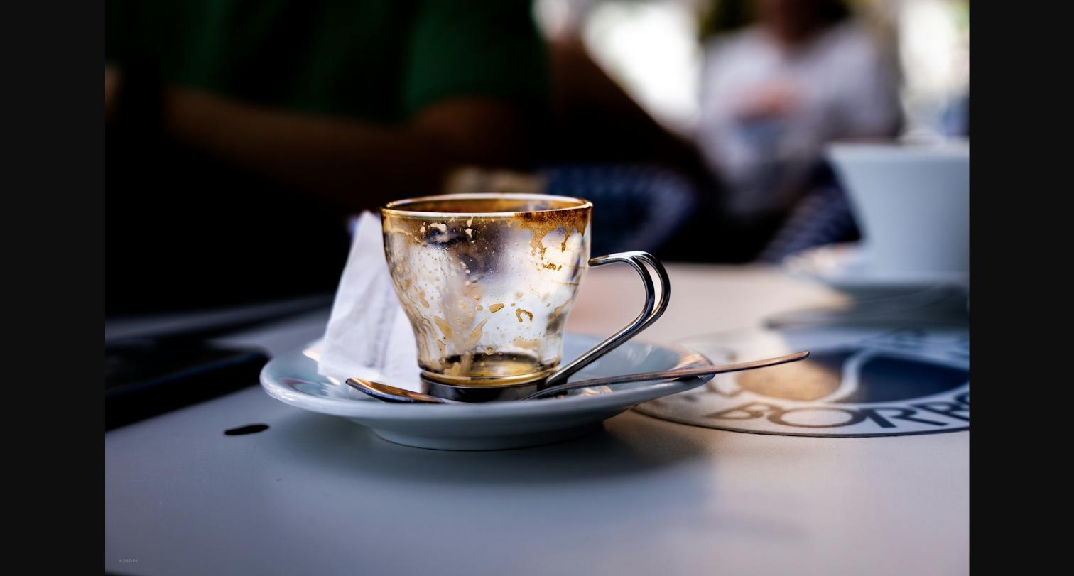 A used espresso cup and saucer left on a café table, out of focus figures in the background.
